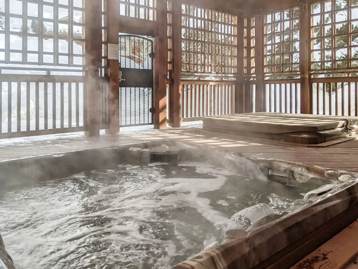 This cozy interior scene centers on a steaming hot tub nestled within a wooden lattice enclosure, with snow gently visible outside. The space invites relaxation and socializing, offering a quintessential après-ski experience in a tranquil setting.