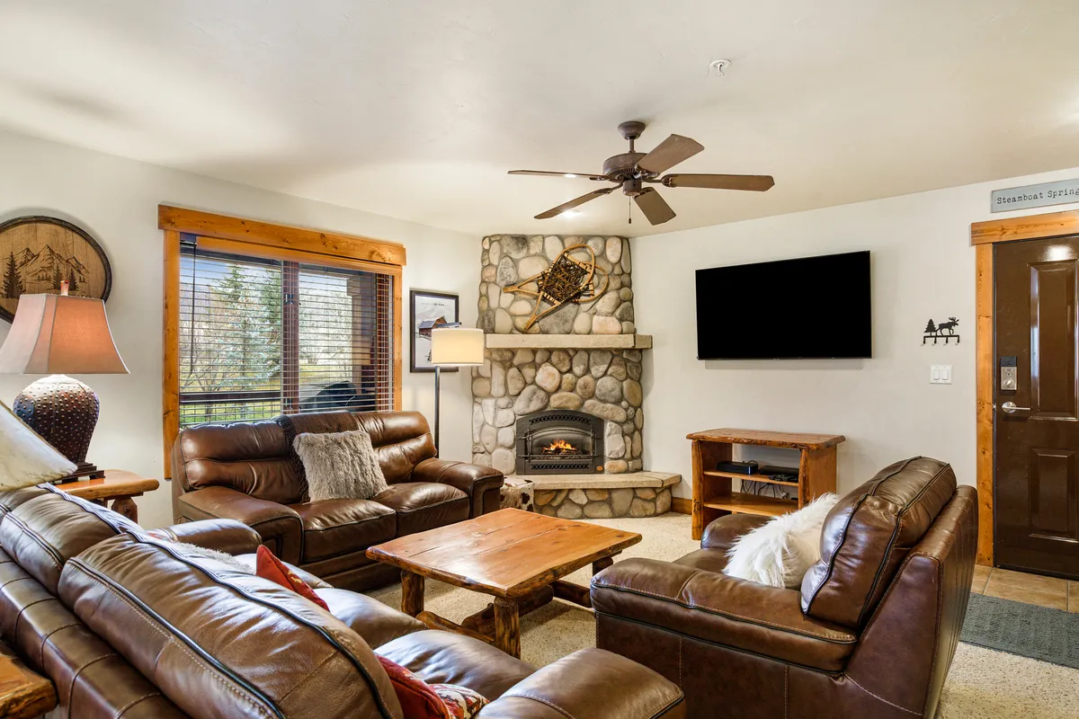 This image captures a cozy living room with a stone fireplace, comfortable leather sofas, a wooden coffee table, and large windows letting in natural light. The setting feels inviting and is perfect for relaxing, socializing, or enjoying a movie night.