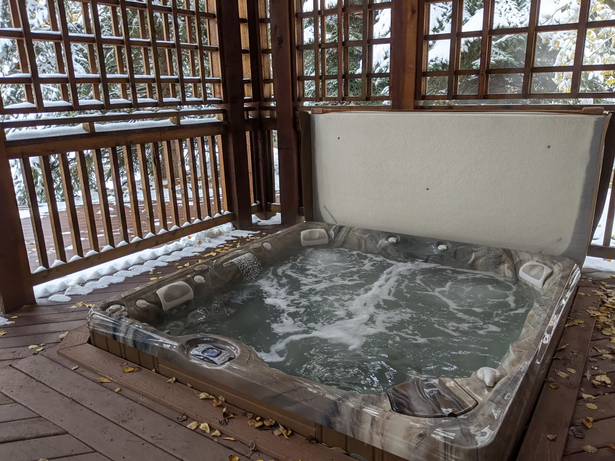 An exterior view of a bubbling hot tub sheltered by a rustic wooden structure, set amidst a scenic winter landscape. People are shown enjoying the warm water, emphasizing both the social and rejuvenating aspects of the amenity.