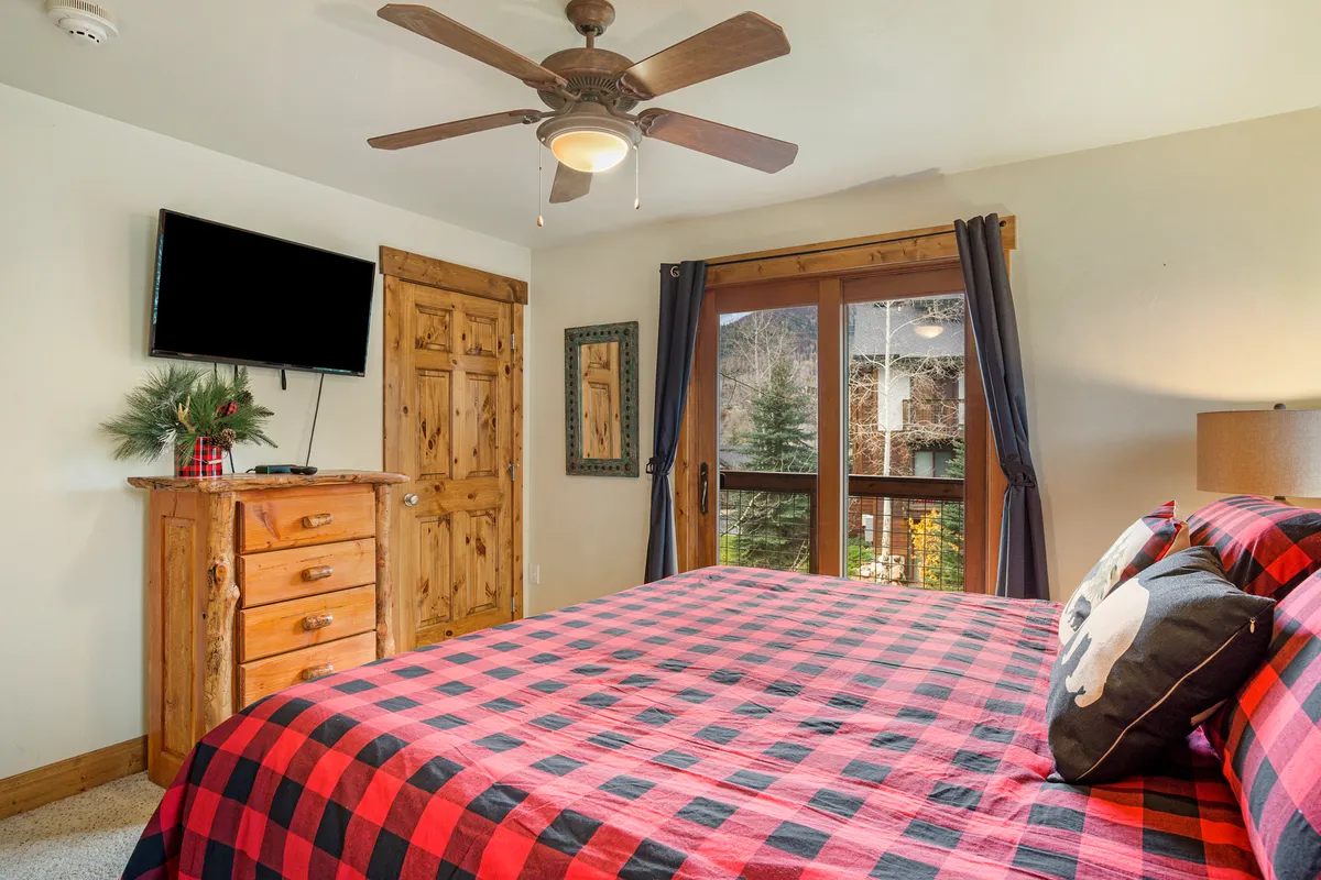 A cozy bedroom scene showcasing a plaid bedspread on a bed, a wooden dresser, and a large window offering views of the outdoors. The room feels comfortable and homey, ideal for relaxing after a day of local exploration or remote work.