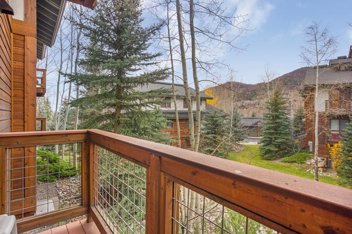 A peaceful covered balcony with a wooden railing overlooking tall trees and distant mountain views. The setting is tranquil, perfect for sipping morning coffee or grilling an evening meal outdoors.