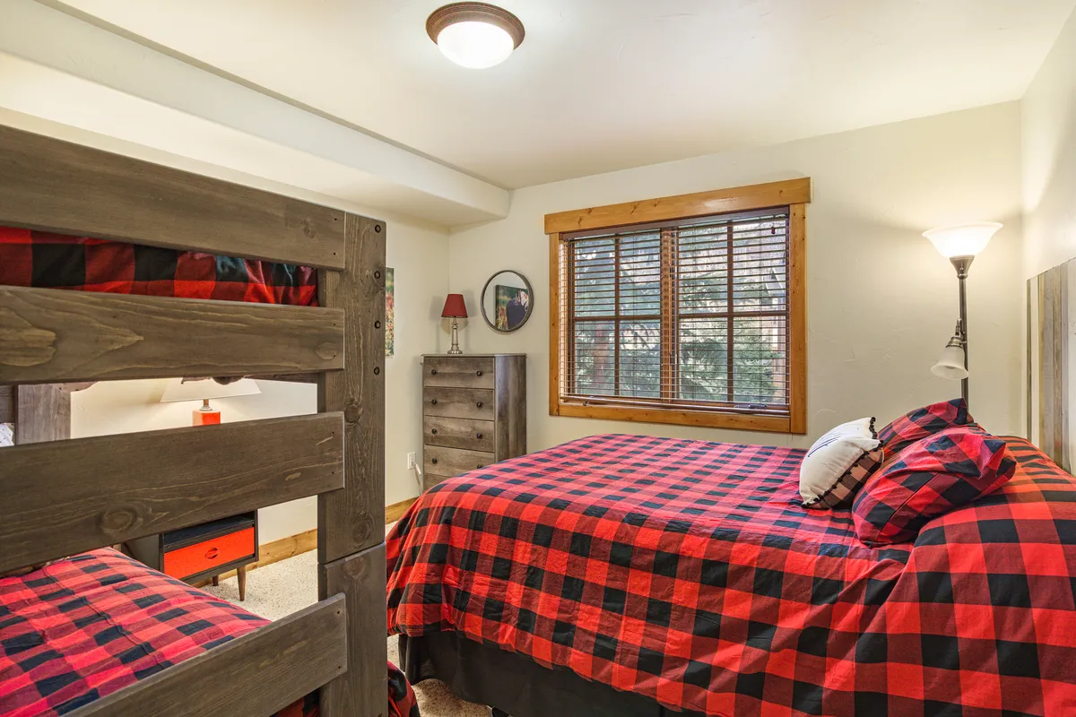 A spacious bedroom featuring both a queen bed and a twin-over-twin bunk bed with red and black checkered bedding. A window with blinds and a reading lamp add functionality, while playful bedding hints at a family-friendly environment.