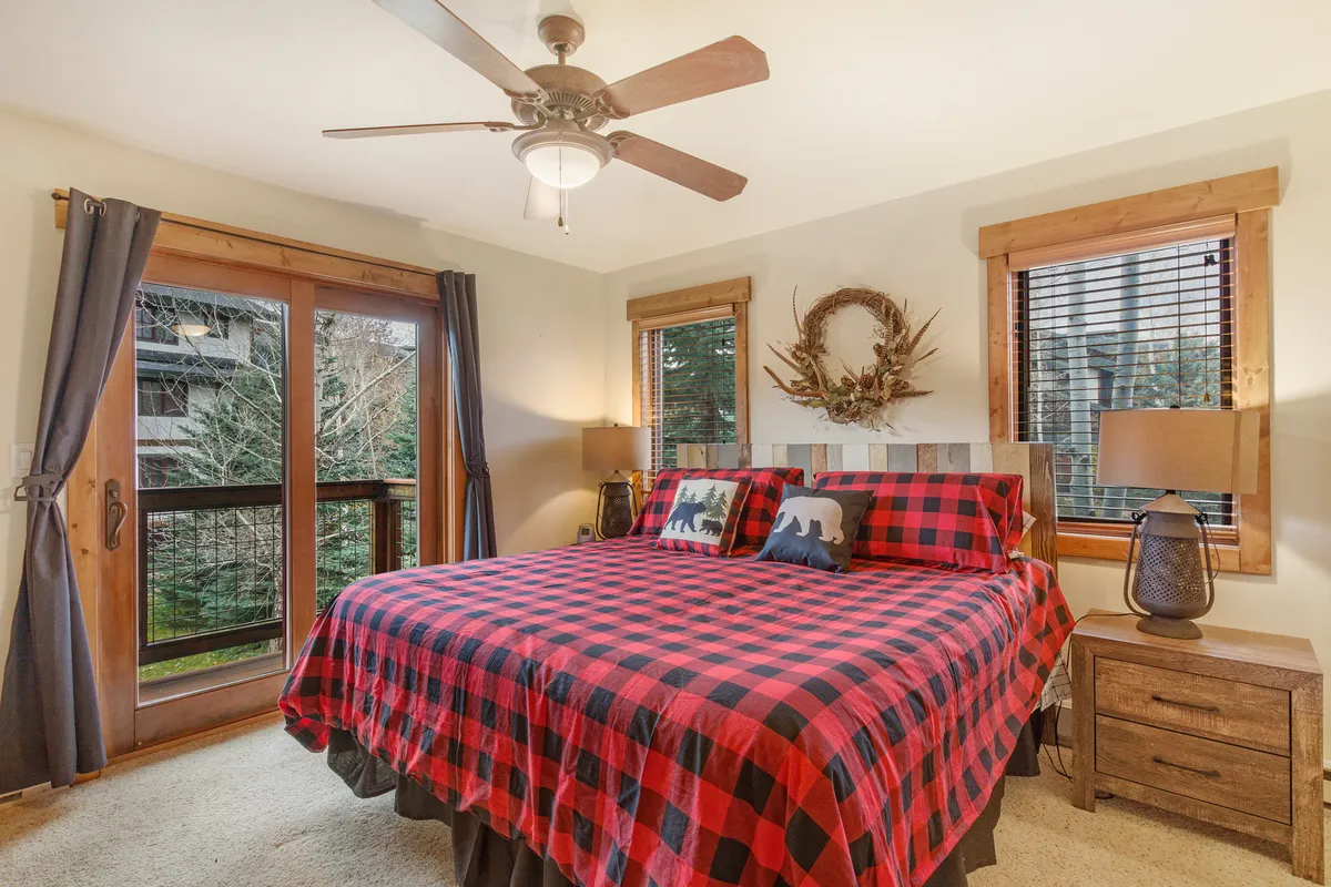 This image showcases a cozy bedroom with plaid bedding, wooden furniture, a decorative wreath on the wall, and large windows that let in abundant natural light. The overall atmosphere is warm and inviting, perfect for restful nights and relaxation.