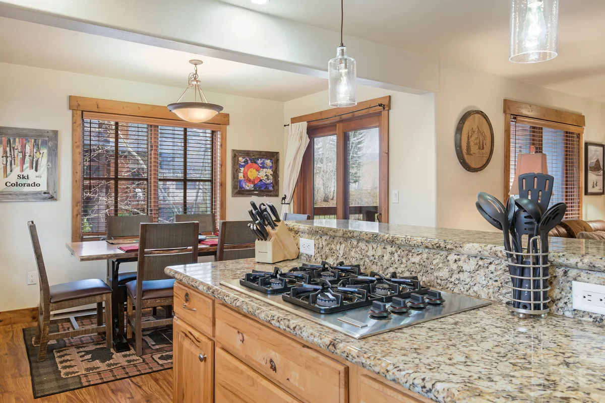 A modern kitchen with a granite countertop, kitchen island stove, adjoining dining area, and a window with a scenic view. The setting is designed for cooking, dining, and socializing in a warm, homey environment.