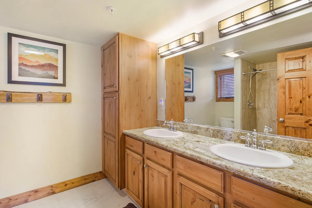 A bright bathroom with a double sink topped by a granite countertop, wooden cabinetry, framed artwork, and a visible shower area. The atmosphere is serene and spa-like, inviting self-care and relaxation.