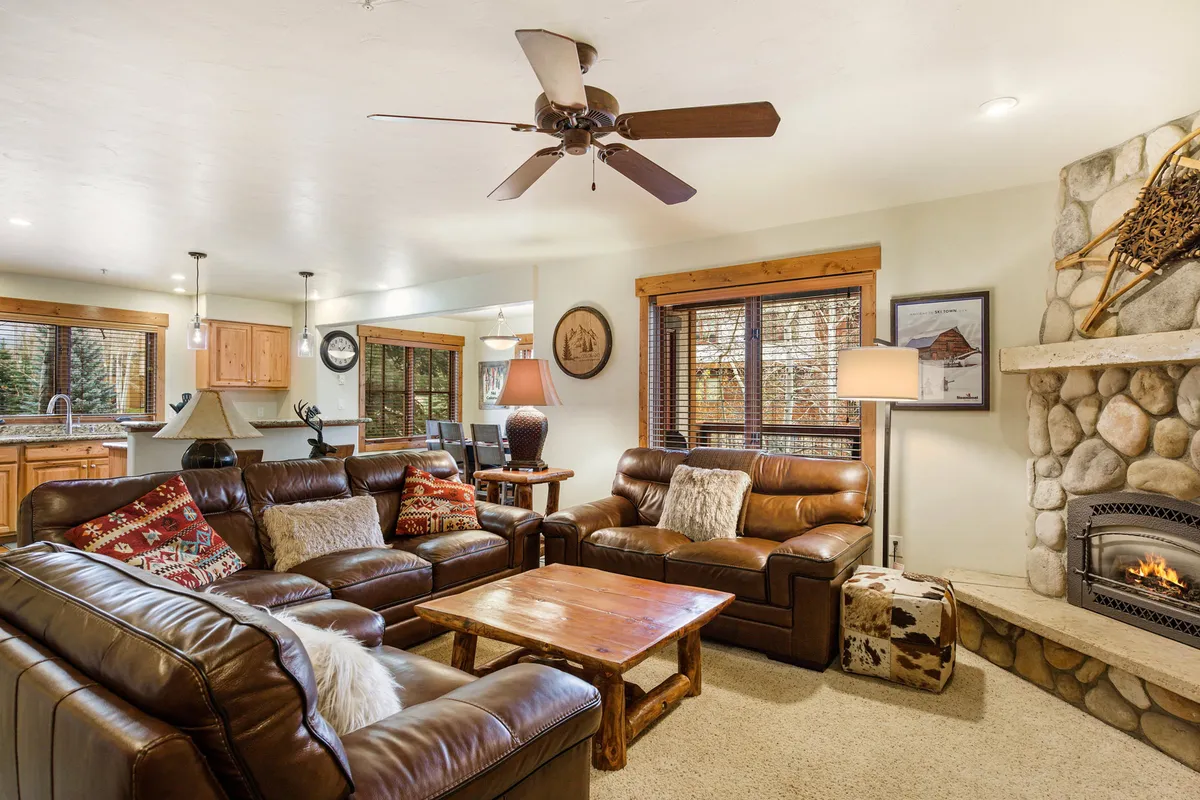 The image showcases a cozy living area featuring plush leather sofas arranged around a rustic stone fireplace, with a wooden coffee table at the center. Large windows frame picturesque views outside, while soft light bathes the room, creating a warm and inviting atmosphere perfect for relaxation or socializing after a day on the slopes.