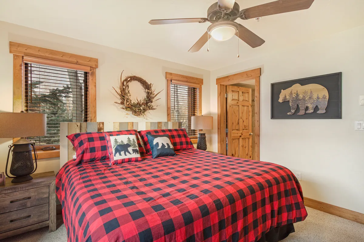A charming bedroom with a checkered comforter, bear-themed pillows, wooden furniture, a decorative wreath, and bear wall art. The room feels intimate and tranquil, ideal for couples seeking privacy and quiet mornings.