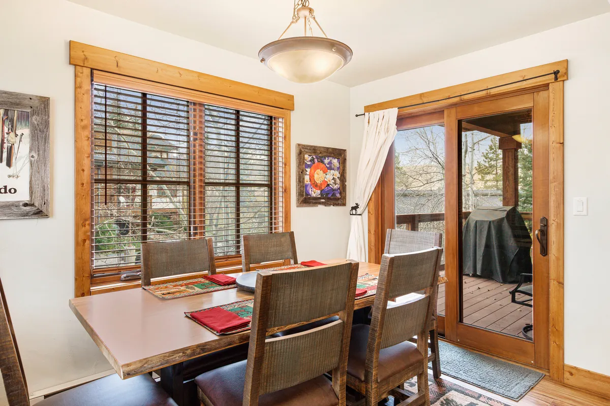 A spacious dining area with a large wooden table, decorative artwork, wooden windows, and a sliding glass door leading to the outdoors. The room is filled with natural light, creating a welcoming space for shared meals or celebrations.